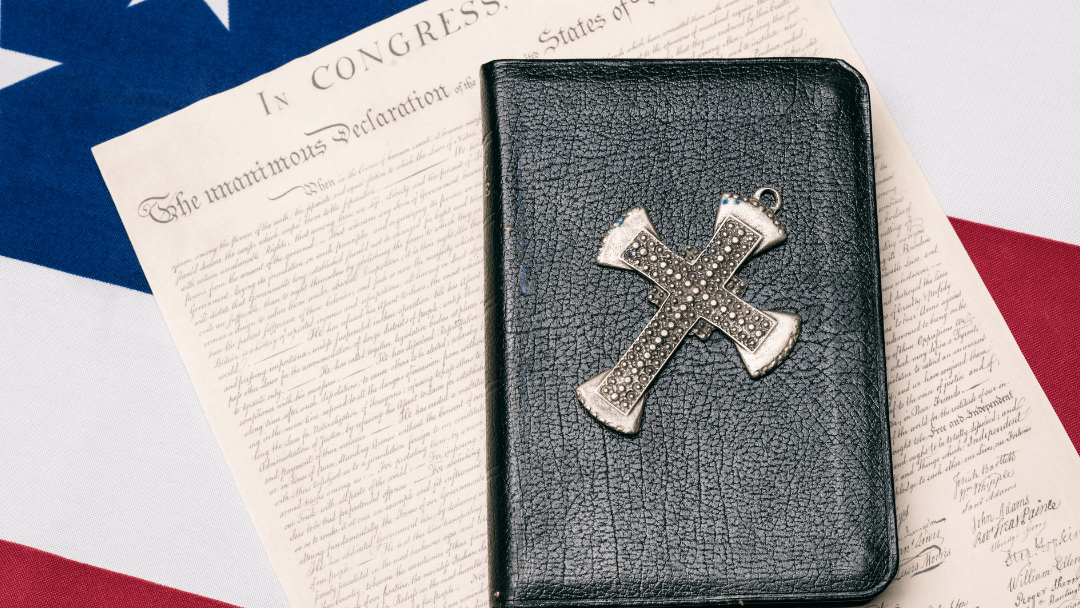 A leather Bible with a jeweled cross pendant resting on the Declaration of Independence over an American flag, symbolizing faith and founding principles.